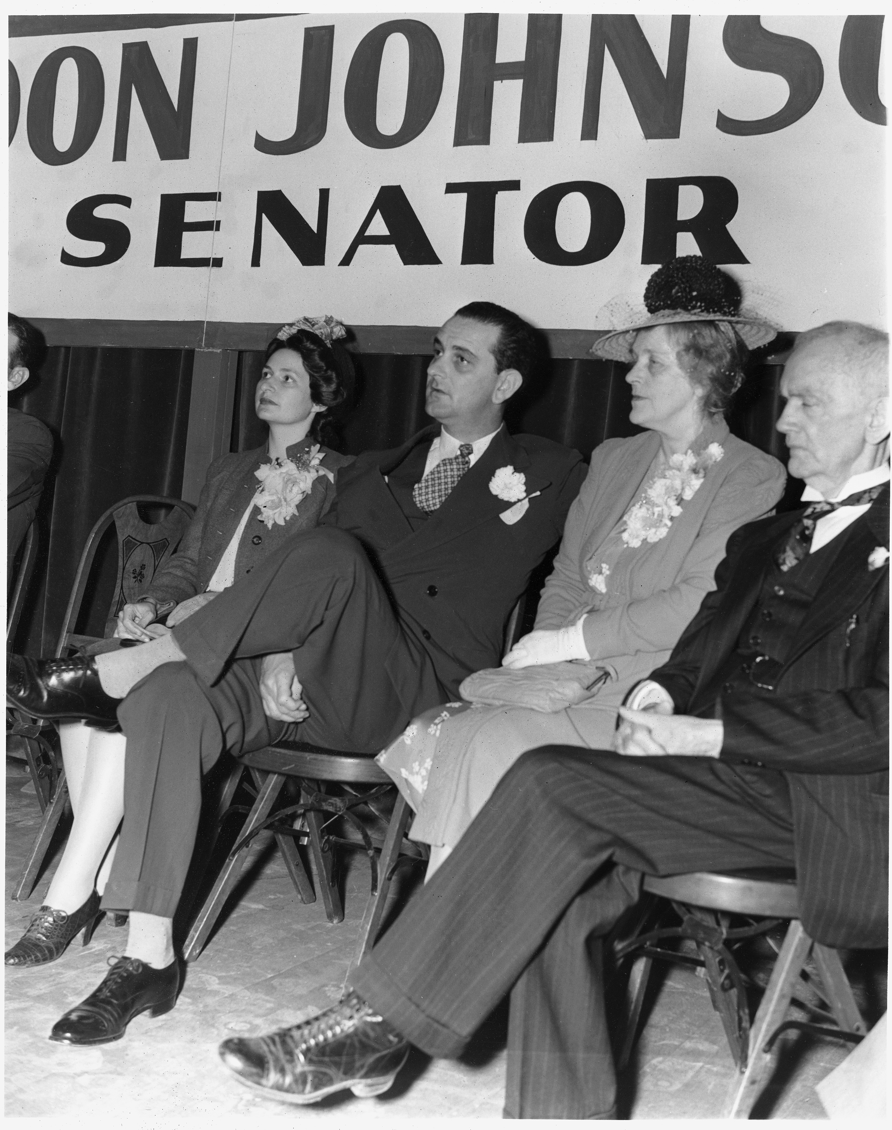 Lady Bird, Lyndon, and Rebekah Johnson sit on a stage with another man under a banner that reads "Lyndon Johnson, Senator"